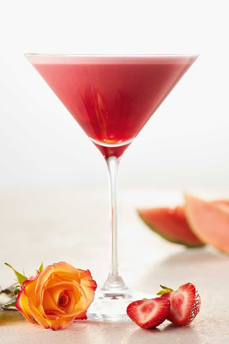 Ein Glas mit Nitro Martini Cocktail, dameben eine Rose und Erdbeeren. 
