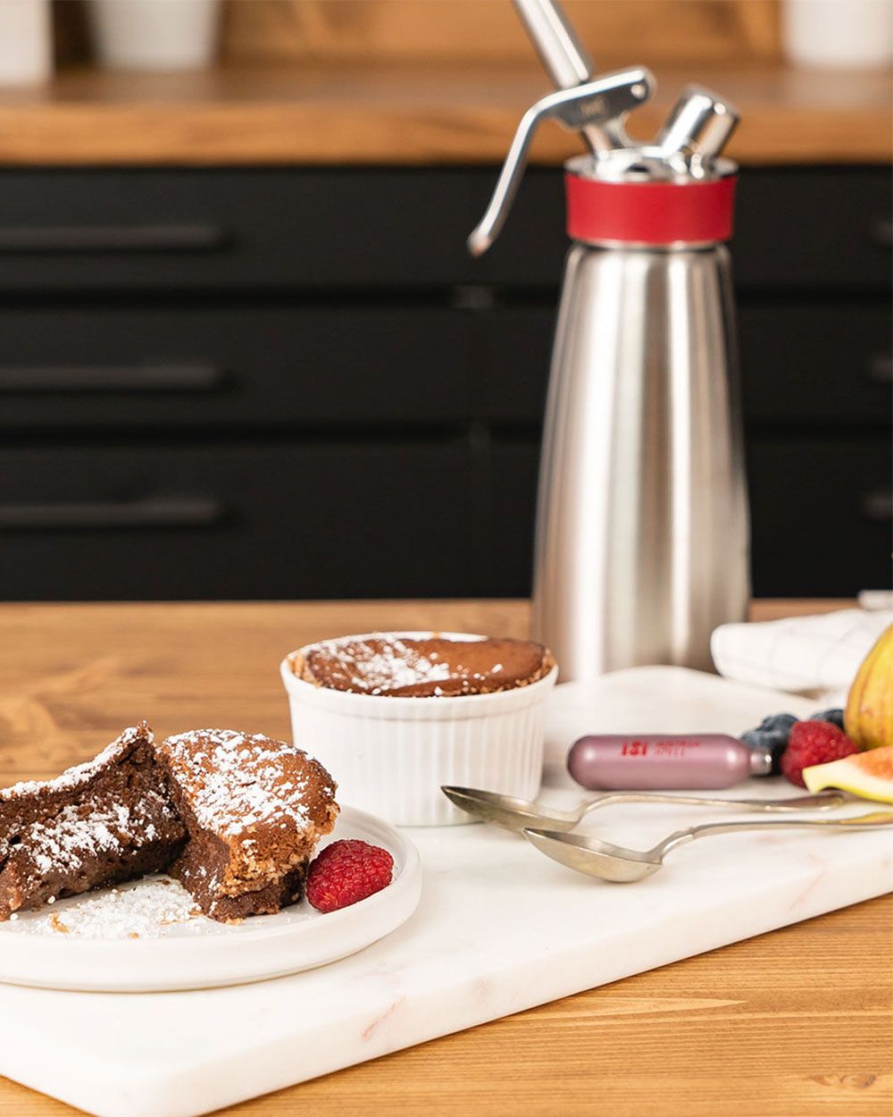 Auf einem Teller liegt ein Schokokuchen, daneben noch ein Schokokuchen in einer kleinen weißen Form. Daneben liegen Löffel und eine pinke iSi Sahnekapsel im Hintergrund steht ein iSi Sahnespender. 