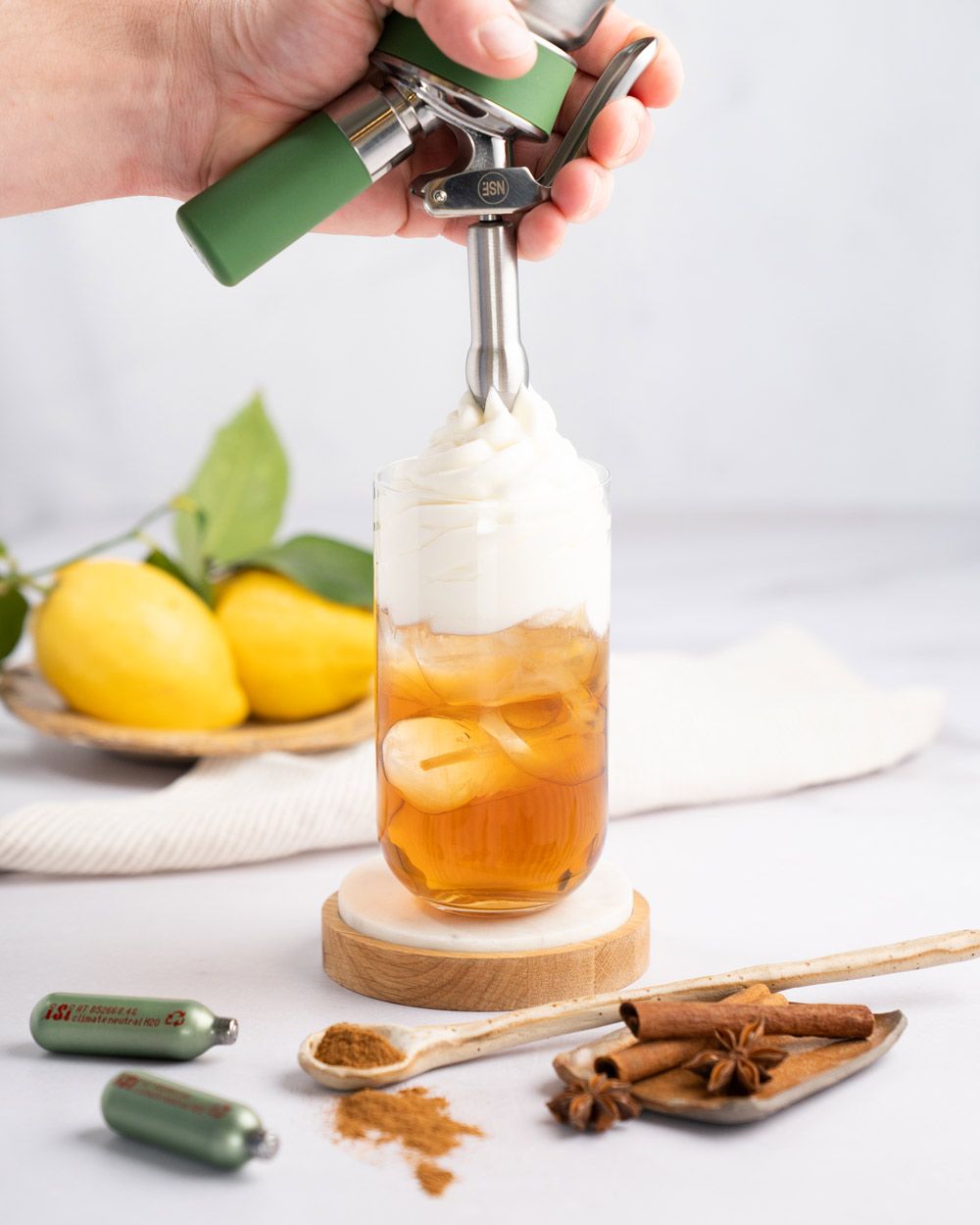 Jemand sprüht Sahne-Topping auf ein Glas mit Eistee, daneben Zimtstangen, iSi Charge Kapseln und Zitronen. 