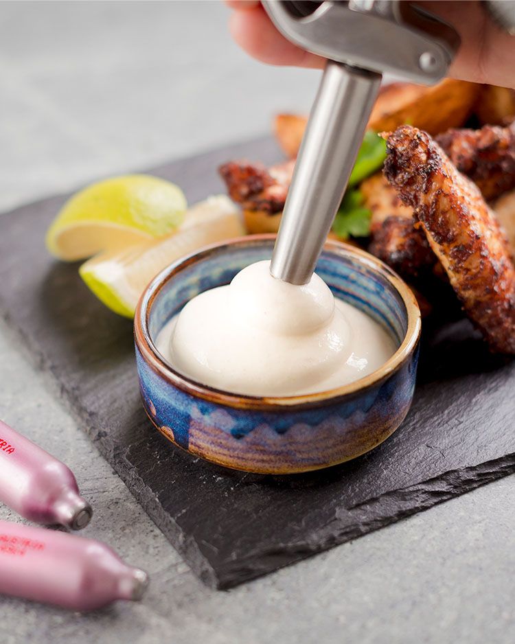 Auf dem Bild wird mit einem iSi Gourmet Whip Aioli in eine kleine Schale auf einem Servierbrett mit Chicken Wings, Kartoffelspalten und Limettenstücken gespritzt.
