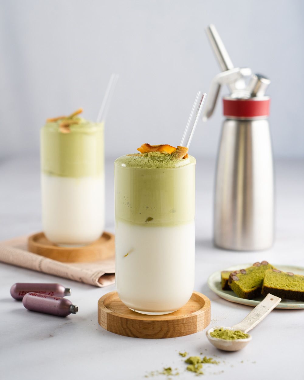 Zwei Milchgetränke mit Matcha-Topping, daneben iSi Charge Kapseln, Matcha-Pulver und eine iSi Gourmet Whip. 