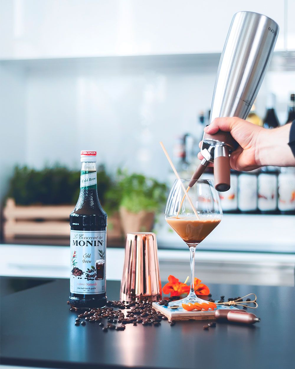 Jemand benutzt einen iSi Nitro, um Nitro Sangria in ein Glas zu srpühen, daneben Kaffeebohnen, iSi Charge Kapseln und Monin Sirup. 