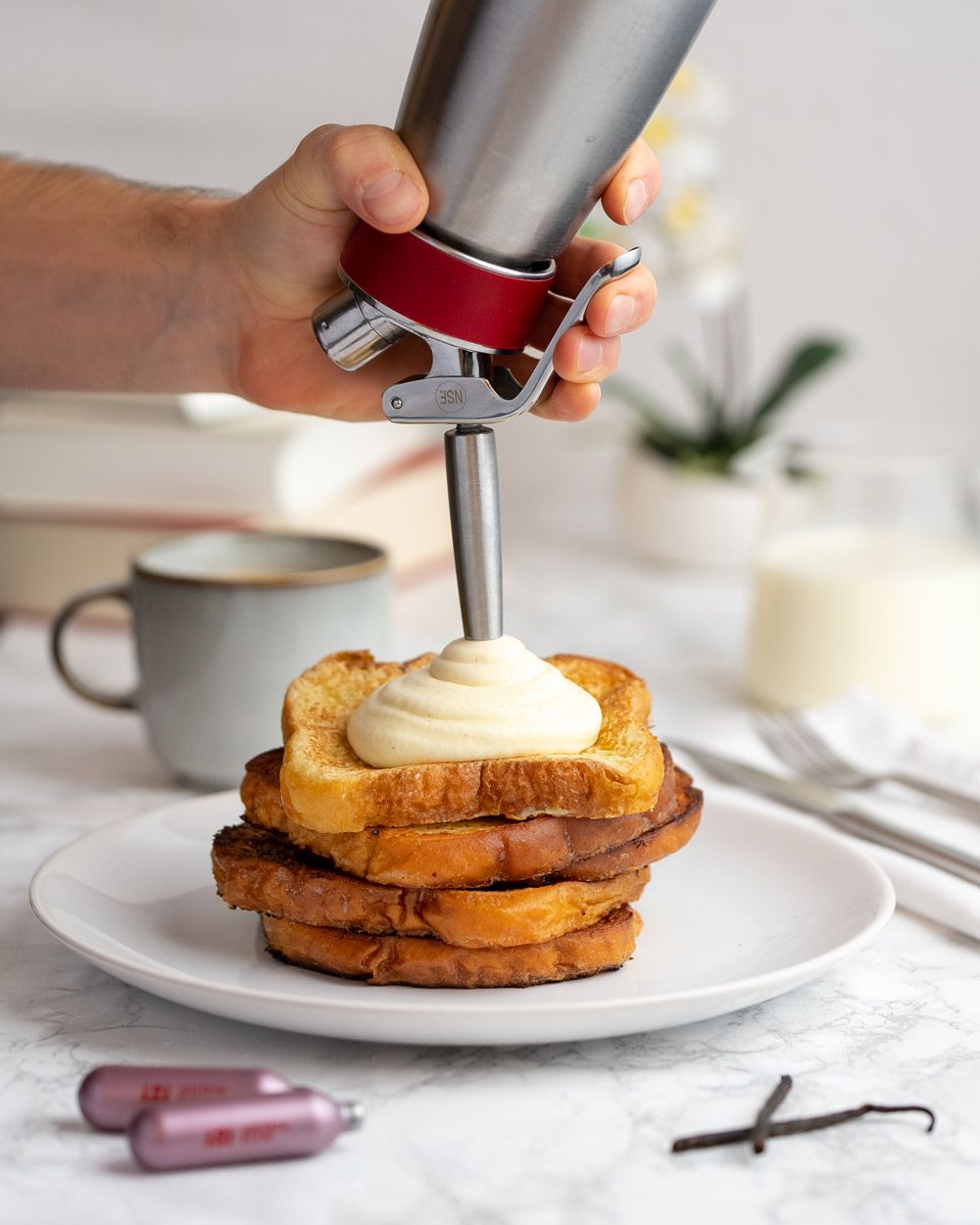 Eine Hand spritzt mit einem iSi Whip cremiges Topping auf gestapelten French Toast, daneben liegen iSi Kapseln und eine Vanilleschote, im Hintergrund stehen eine Tasse Kaffee und ein Glas Milch.