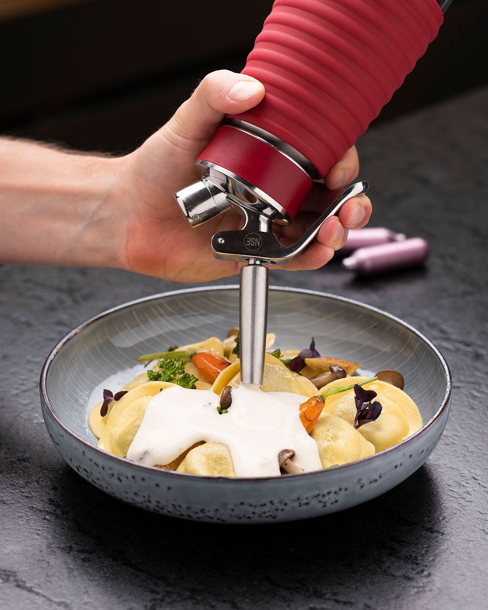 Jemand sprüht mit einem iSi Gourmet Whip Pasta Sauce auf einen Teller Pasta, daneben iSi Charge Kapseln. 