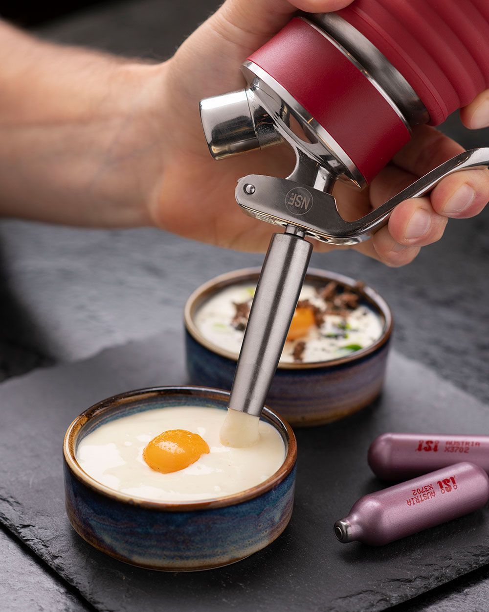 Jemand sprüht mit einem iSi Gourmet Whip Kartoffel-Espuma in eine Schüssel, daneben iSi Charge Kapseln und eine weitere Schüssel mit Espuma.