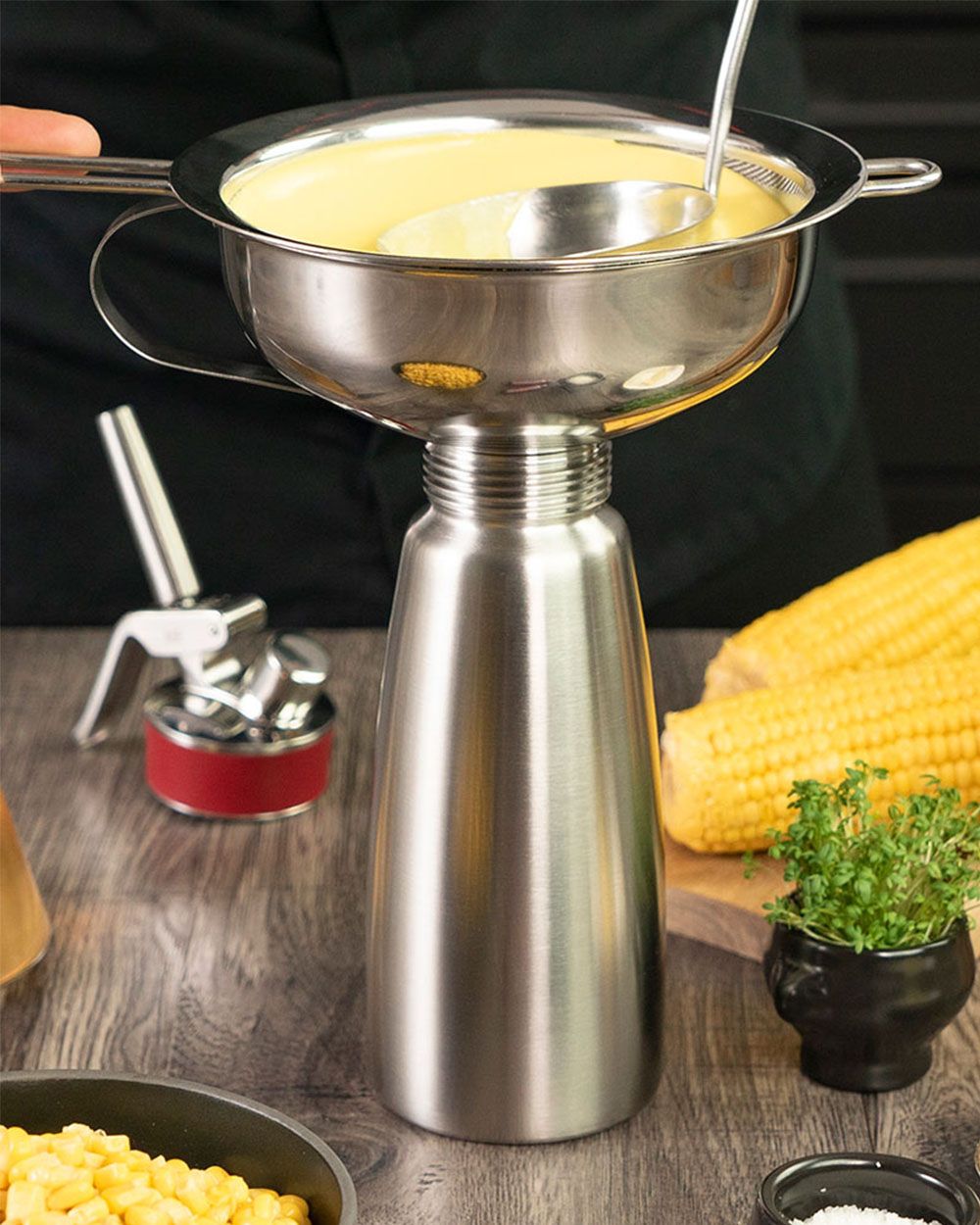 Eine Kelle mit cremiger Maissuppe wird durch einen Trichter in einen iSi Whip gefüllt, während daneben Maiskolben, Kräuter und eine iSi Kapselhalterung auf dem Tisch liegen.