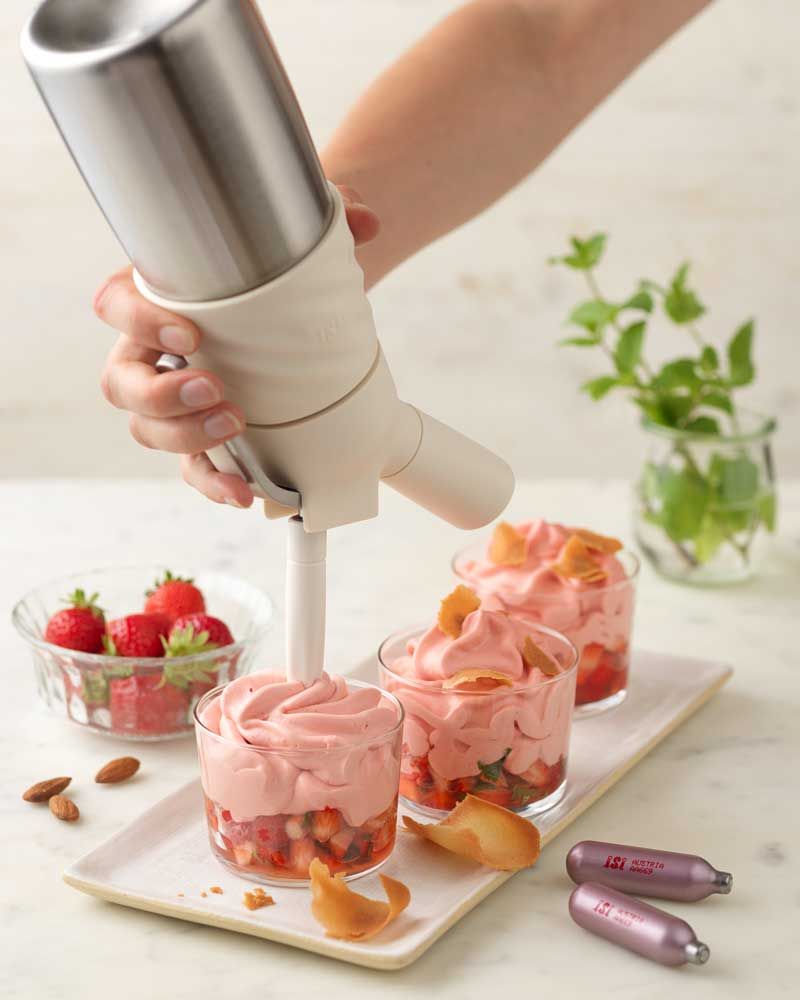 Jemand nutzt einen iSi Creative Whip um Erdbeermousse in Gläser zu füllen. Daneben Erdbeeren und zwei iSi Charge Kapseln.