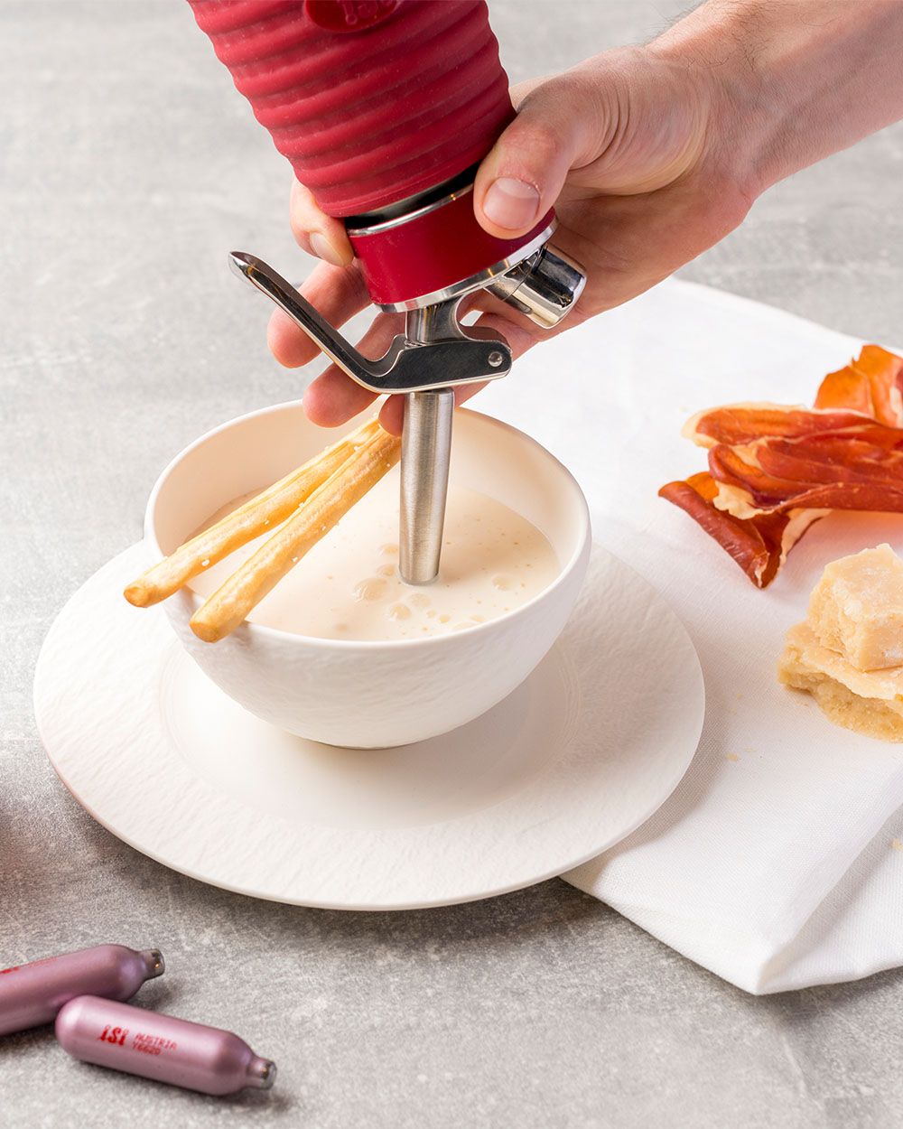 Jemand sprüht mit einem iSi Gourmet Whip Parmesan Espuma in eine Schüssel, daneben Prosciutto, Parmesan und zwei iSi Charge Kapseln. 