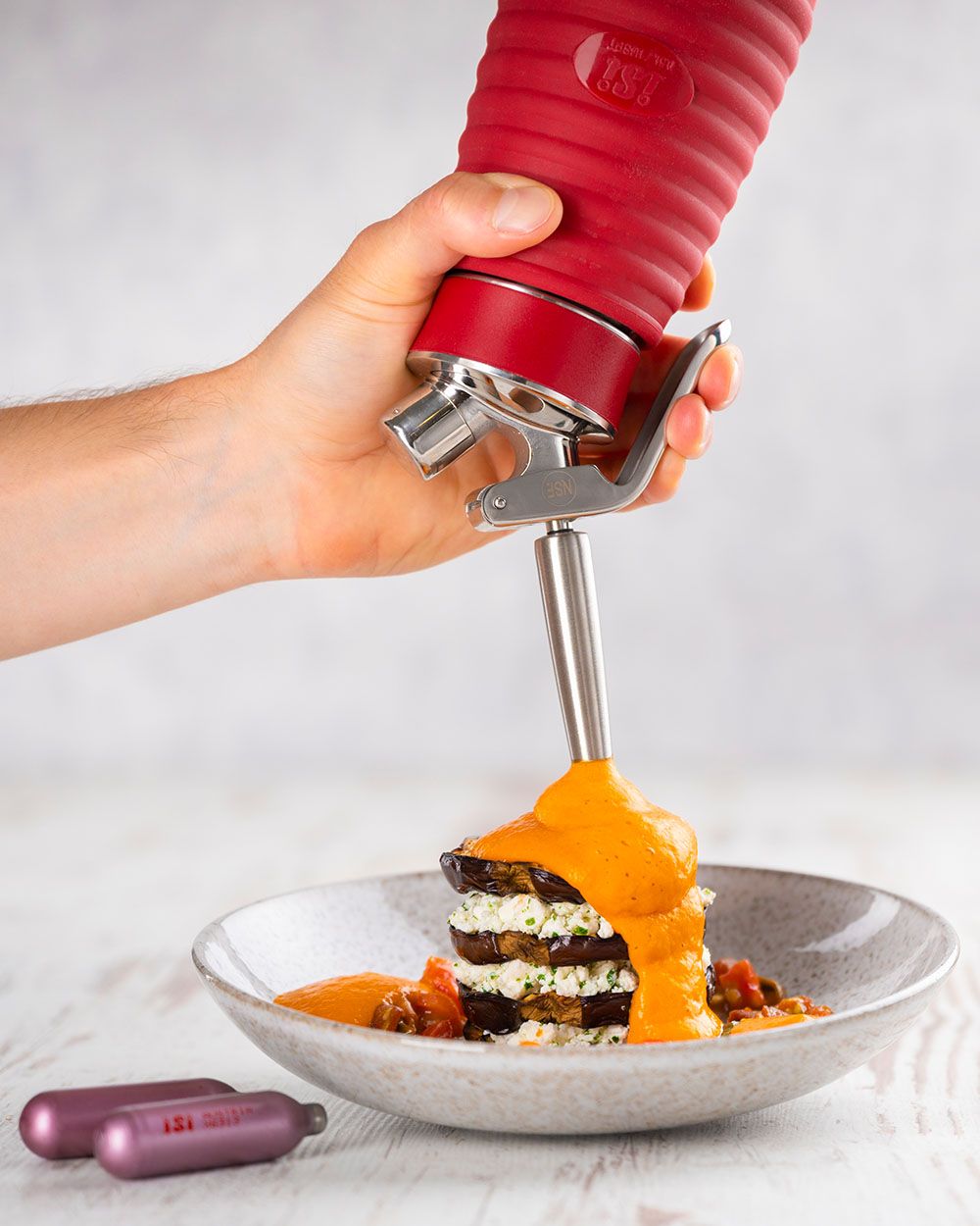 Jemand sprüht mit eine iSi Gourmet Whip Tomaten-Parmesan-Sauce auf einen Teller, daneben iSi Charge Kapseln. 