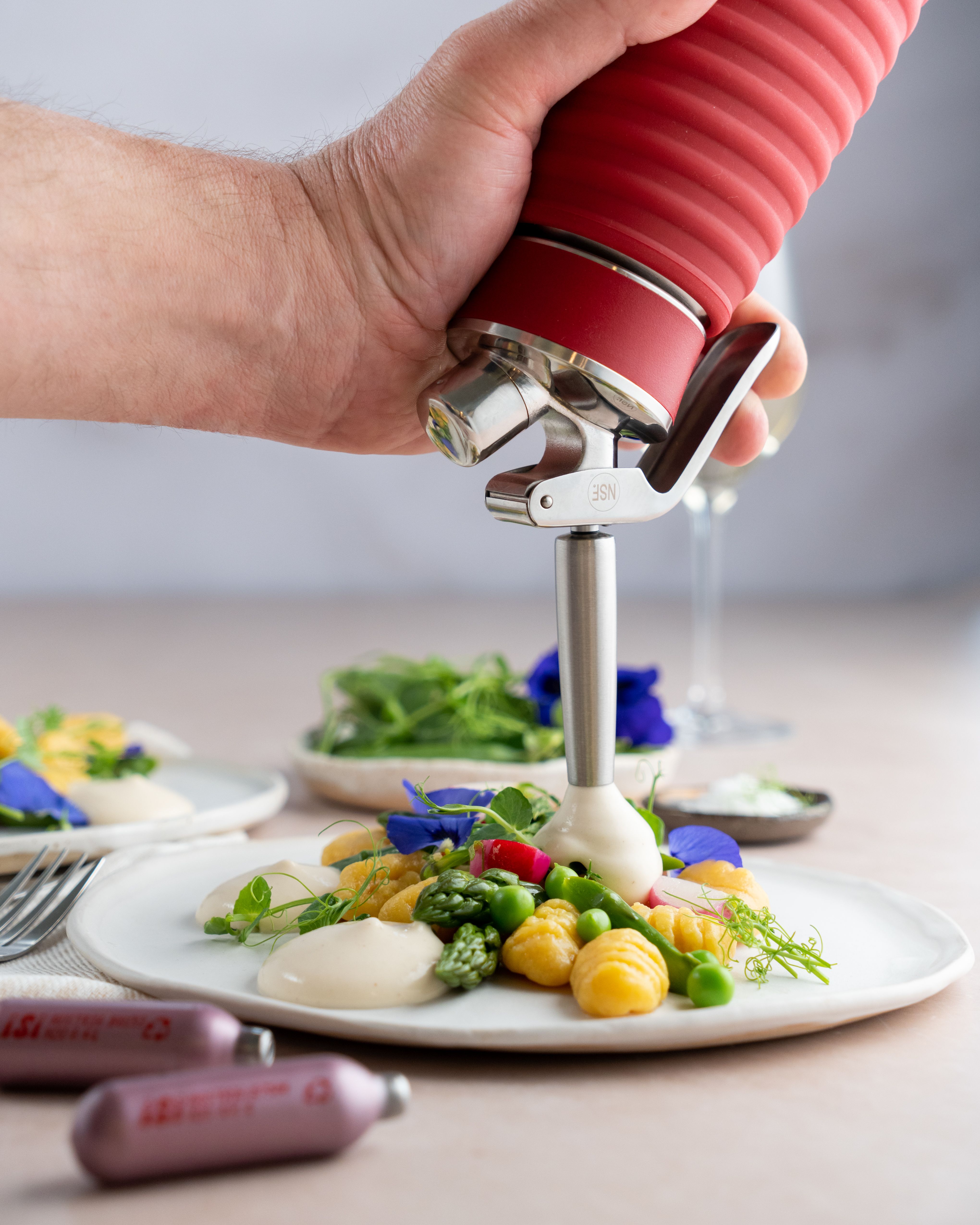 Jemand sprüht mit einem iSi Gourmet Whip Spargel-Espuma auf einen Teller mit Gnocchi und Gemüse, daneben iSi Charge Kapseln.