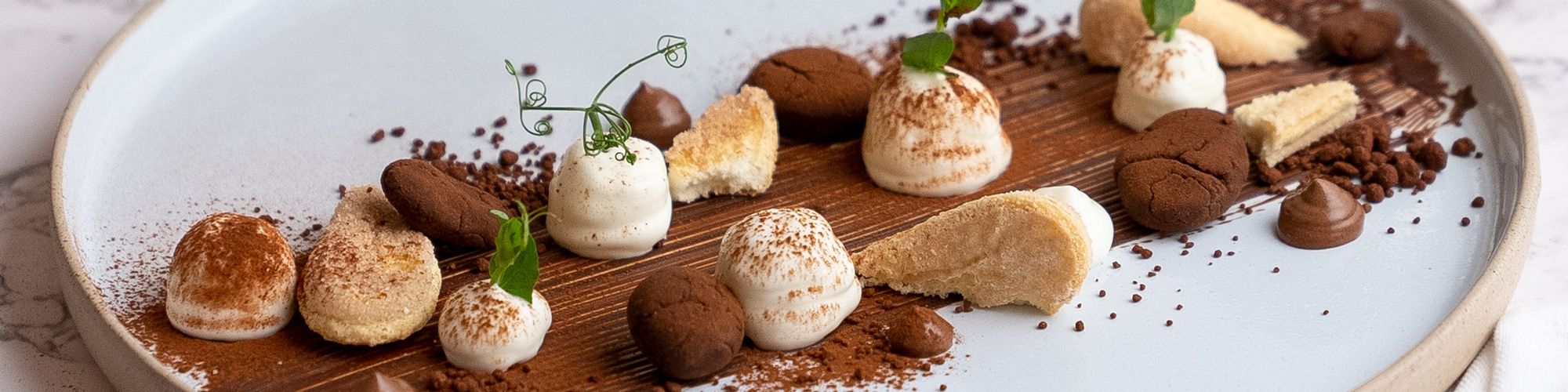 Ein Dessertteller ist kunstvoll mit kleinen Cremehäubchen, Schokoladenstückchen, Gebäck und Kakaopulver angerichtet.
