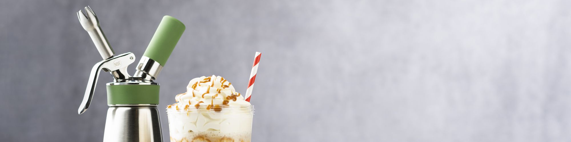 Ein iSi Green Whip steht neben einem Glas mit Sahnehaube, Karamellstückchen und einem rot-weißen Strohhalm.