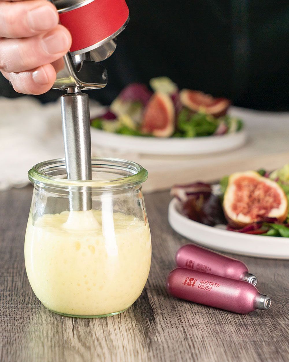 Eine Hand spritzt mit einem iSi Whip gelbes Schaum-Dressing in ein Glas, daneben liegen iSi Kapseln und im Hintergrund steht ein Teller mit frischem Salat und Feigen.