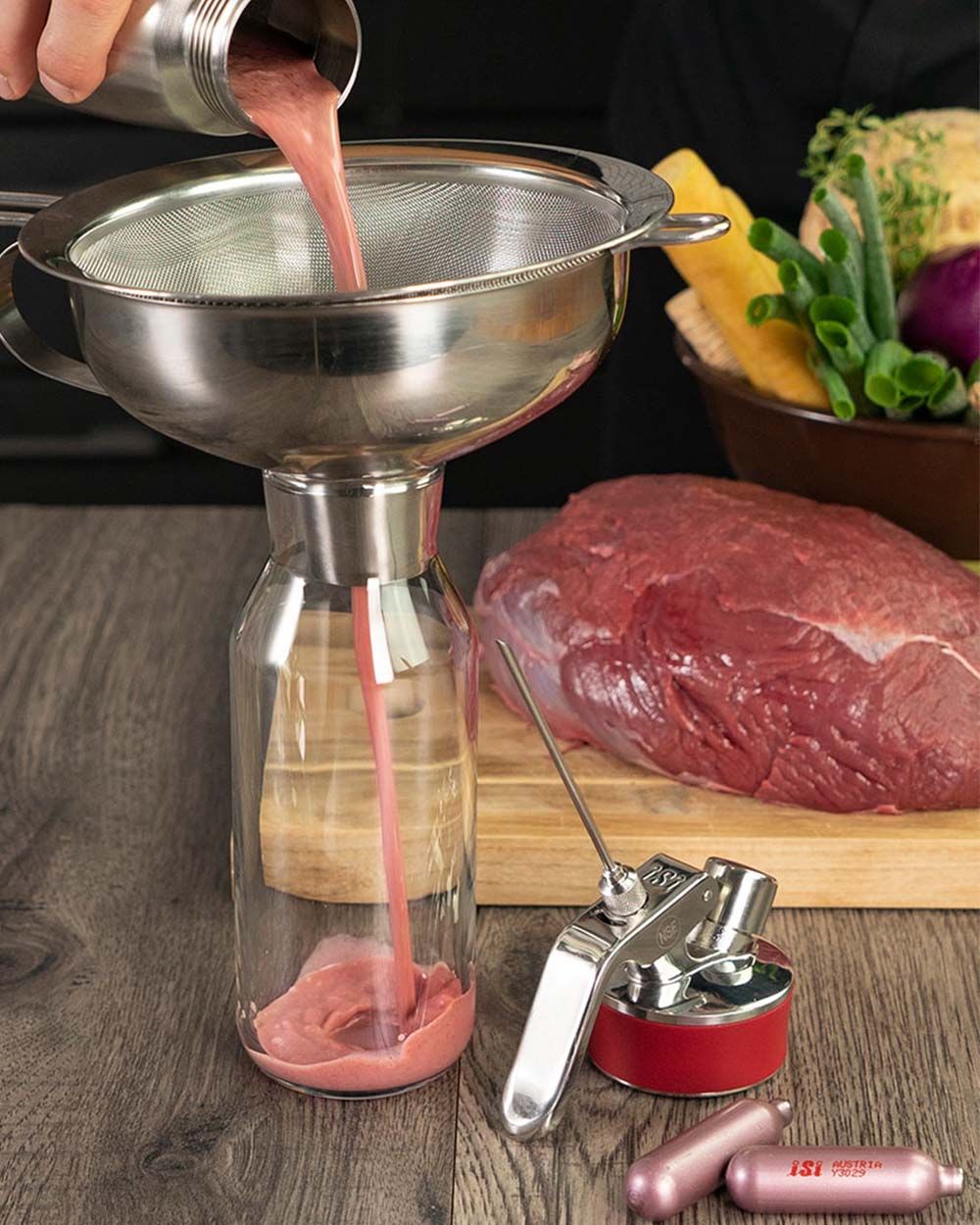 Jemand gießt rosafarbene Sauce aus einem iSi Gourmet Whip in einen Krug, im Hintergrund liegt ein großes Stück Fleisch. 