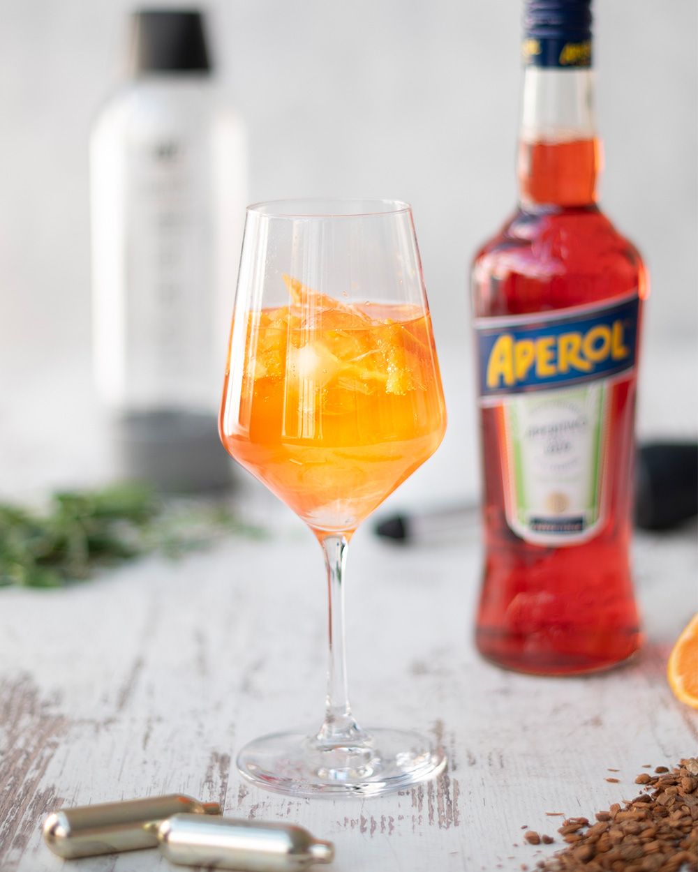 Ein Glas mit Aperol Spritz, im Hintergrund eine Flasche Aperol, davor iSi Charge Kapseln. 