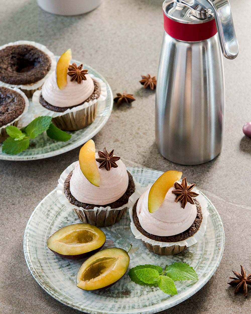 Zwei Teller mit Mohn-Pflaumen-Cupcakes, daneben Pflaumen und ein iSi Gourmet Whip. 