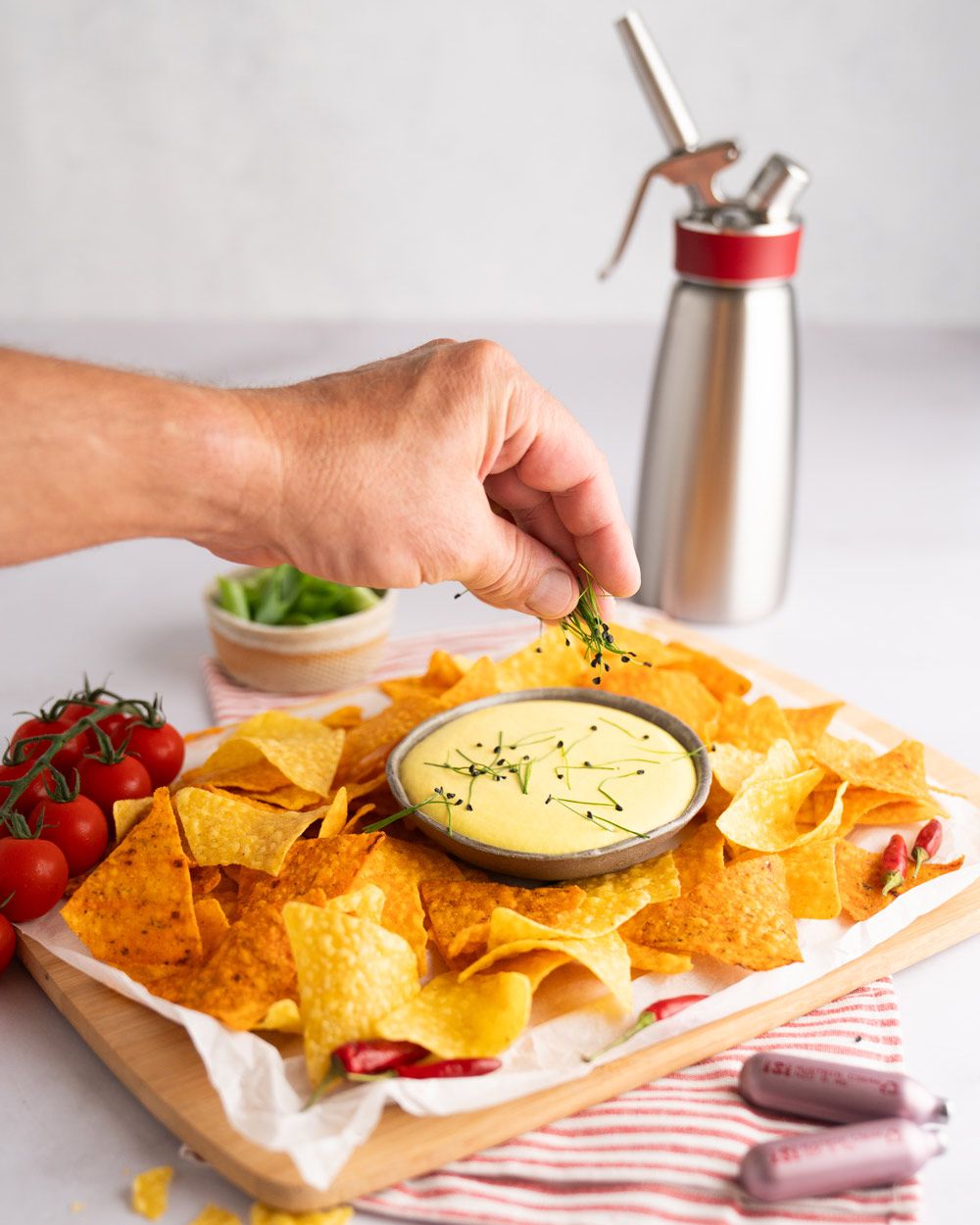 Jemand gibt Kräuter auf eine Schüssel mit Käse-Espuma, daneben Nachos. Davor zwei iSi Charge Kapseln, im Hintergrund ein iSi  Gourmet Whip. 