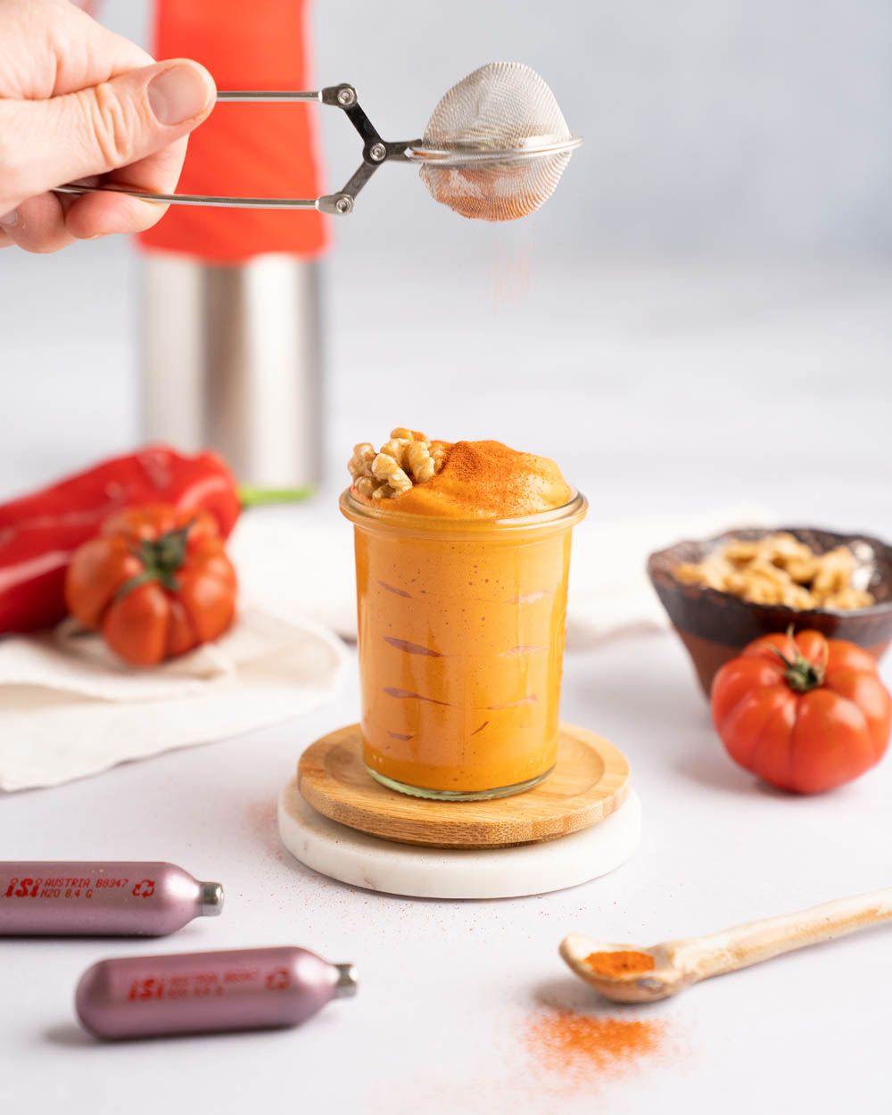 Jemand gibt Gewürze auf ein Glas mit Muhammara Dip, daneben iSi Charge Kapseln, Gewürze, Tomaten und Walnüsse. Im Hintergrund ein iSi Creative Whip. 