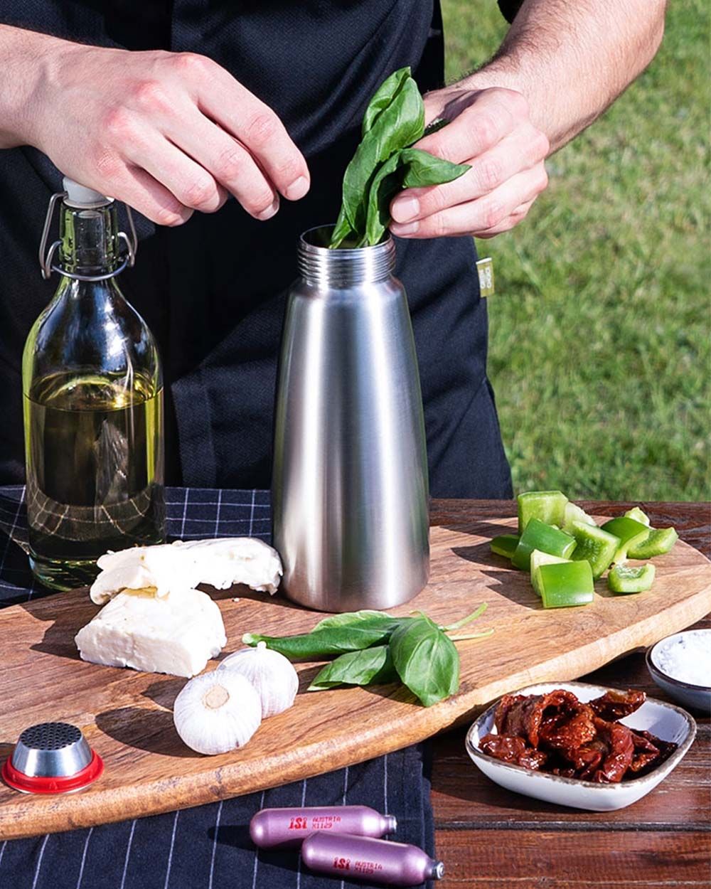 Jemand gibt Basilikum in eine iSi Gourmet Whip, daneben weitere Gewürze, Halloumi, Knoblauch, getrocknete Tomaten, Öl und iSi Charge Kapseln. 