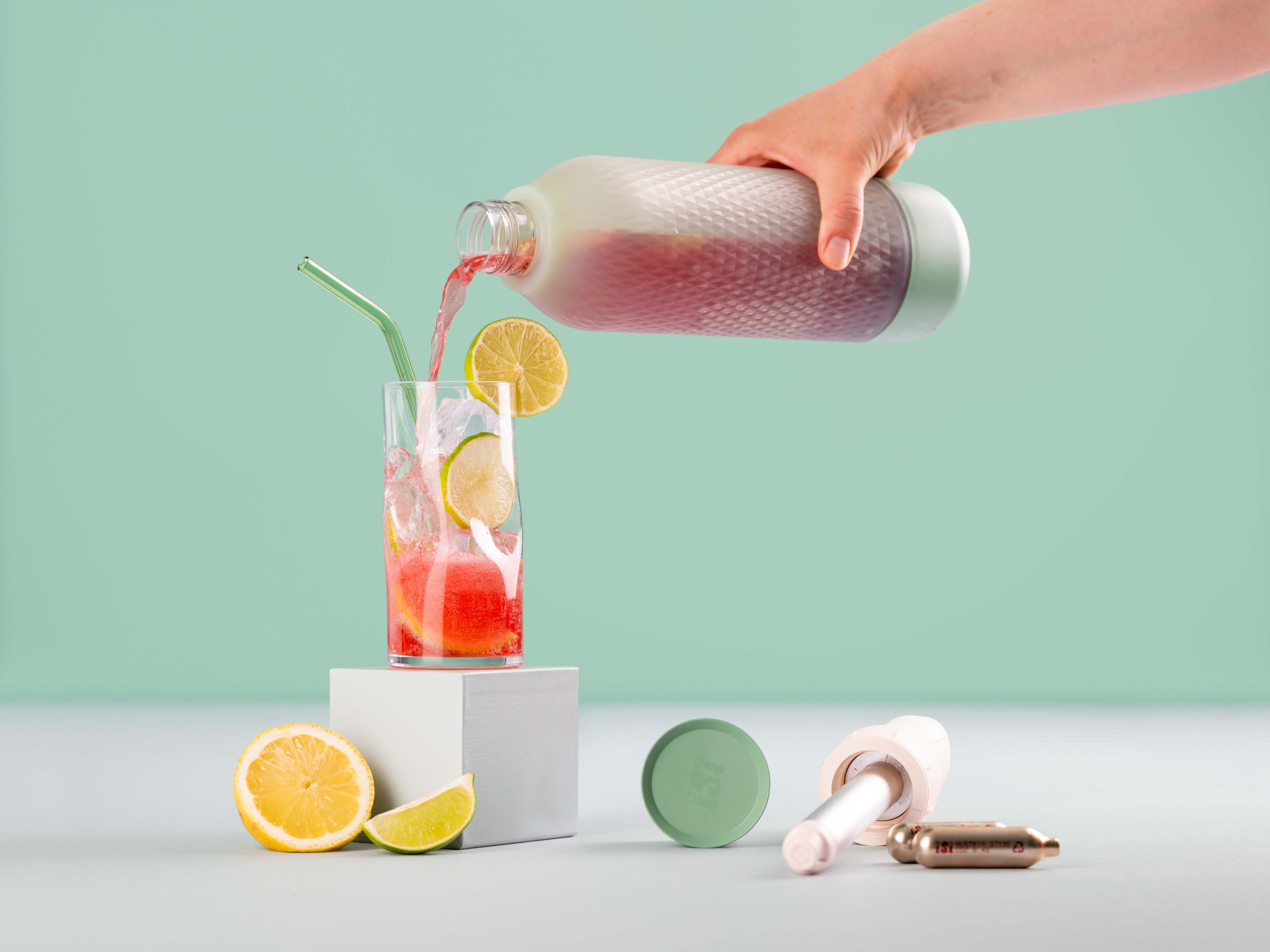 Jemand gießt Cranberry Ginger Lemonade aus einem iSi Twist'n Sparkle in ein Glas mit Eis und Zitronen, daneben weitere Zitronen, ein Twist'n Sparkle Aufsatz und iSi Charge Kapseln.