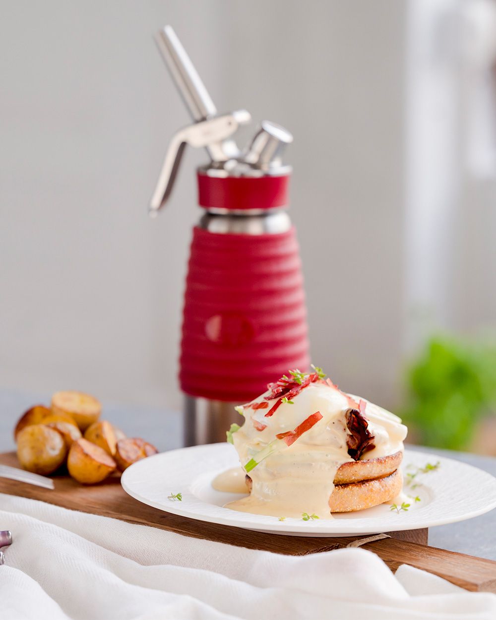 Eggs Benedict mit Hollandaise Sauce, im Hintergrund Kartoffelscheiben und ein iSi Gourmet Whip. 