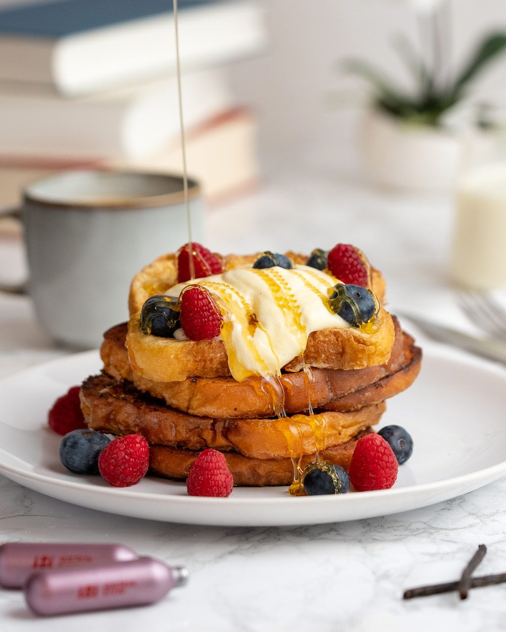 Gestapelter French Toast mit Beeren und cremigem Topping wird mit Honig übergossen, daneben liegen iSi Kapseln und eine Vanilleschote, im Hintergrund stehen Bücher, eine Tasse Kaffee und ein Glas Milch.
