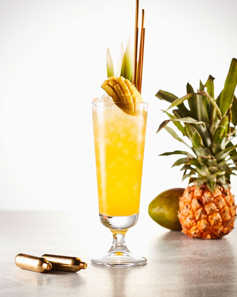Ein gelbes Getränk – Bangkok Limonade – daneben zwei goldene iSi Kapseln, eine Ananas und eine Mango. 