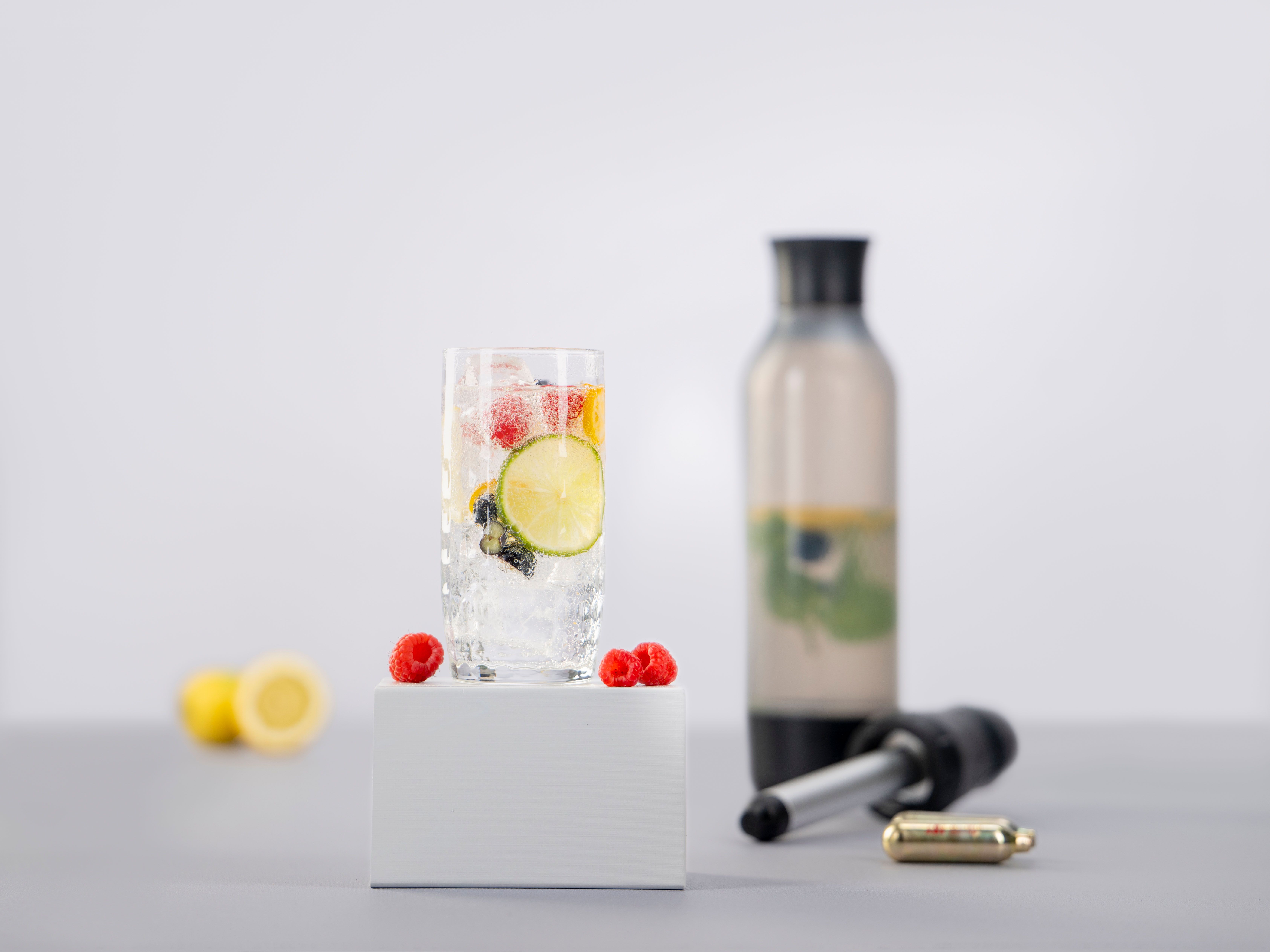Ein Glas mit Sparkled Infused Wasser und Früchten, daneben weitere Früchte, ein iSi Twist'n Sparkle und iSi Charge Kapseln.