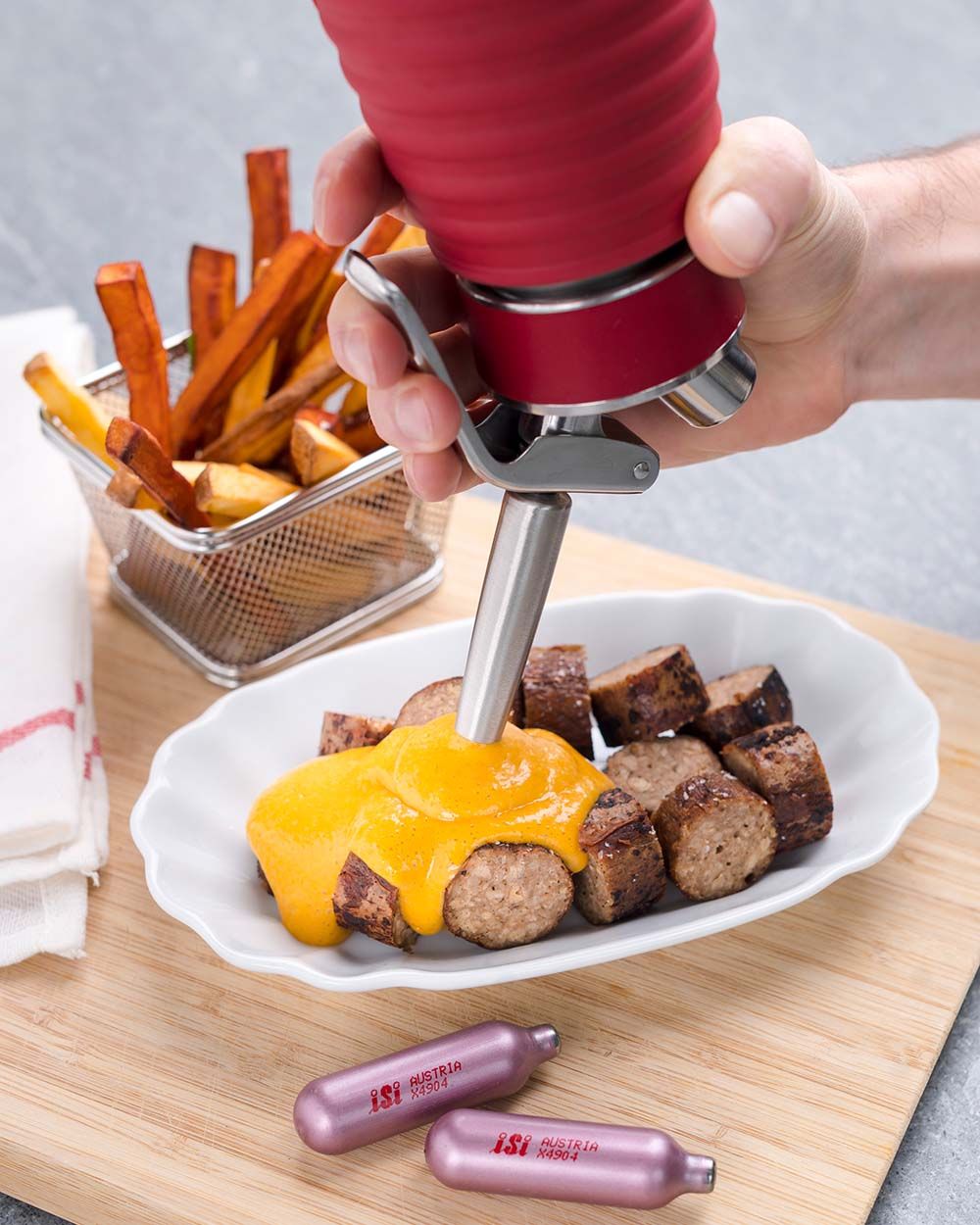 Jemand sprüht vegane Curry Sauce auf einen Teller mit Würsten, daneben Pommes und iSi Charge Kapseln.