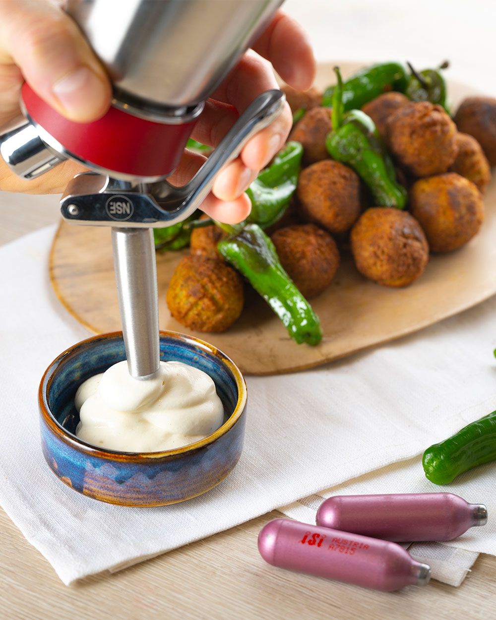 Ein Teller mit Falafeln und Jalapenos, daneben eine Schüssel in die jemand mit einem iSi Creative Whip Aioli sprüht.