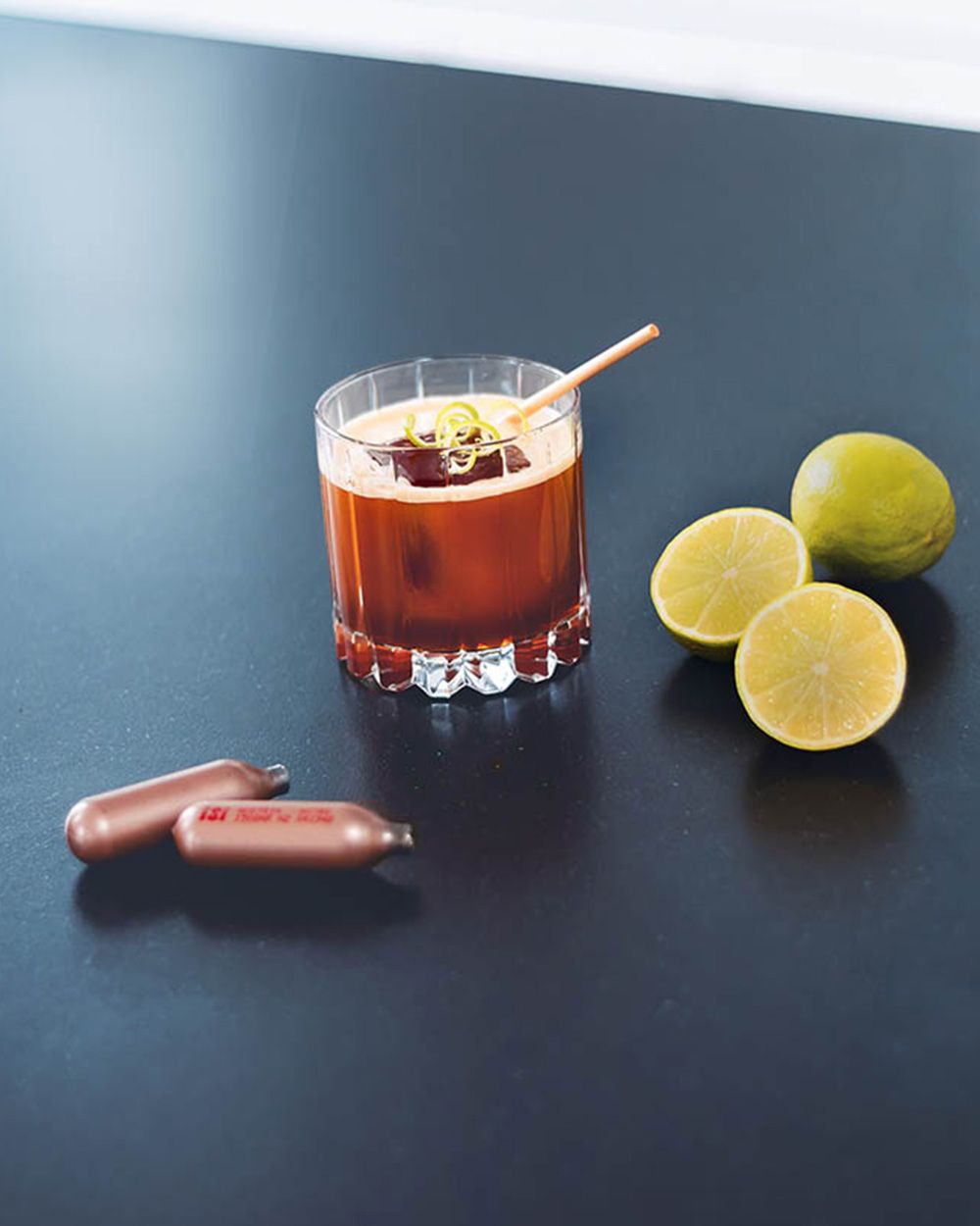 Ein Glas mit einem Kaffee-Gin-Getränk, daneben iSi Charge Kapseln und zwei Limetten. 