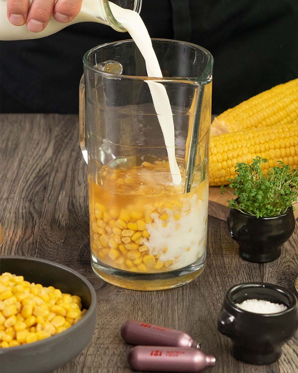 In einen Krug mit Maiskörnern wird Milch gegossen, während daneben frische Maiskolben, Kräuter, Salz, eine Schale mit Mais und iSi Kapseln auf dem Tisch liegen.