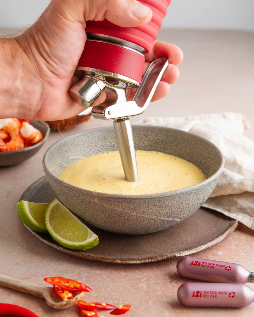 Jemand sprüht mit einem iSi Gourmet Whip thailändische Kokos-Suppe in einen Teller, daneben iSi Charge Kapseln. 