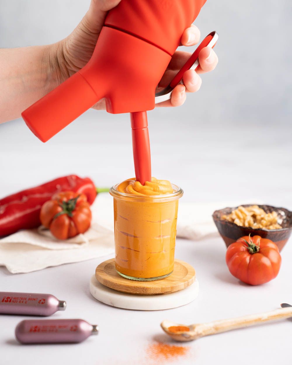 Jemand sprüht mit einem iSi Creative Whip Muhammara Dip in ein Glas, daneben iSi Charge Kapseln, Gewürze, Tomaten und Paprika.