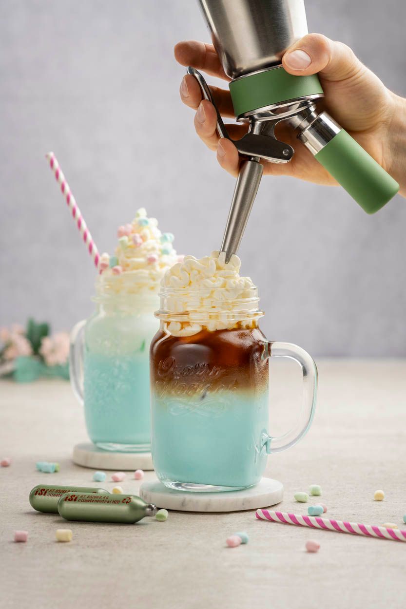 Jemand sprüht auf ein blaues Frappe-Getränk mit einem iSi Green Whip Sahneschaum, im Hintergrund steht ein fertiger Bubblegum Frappe. 