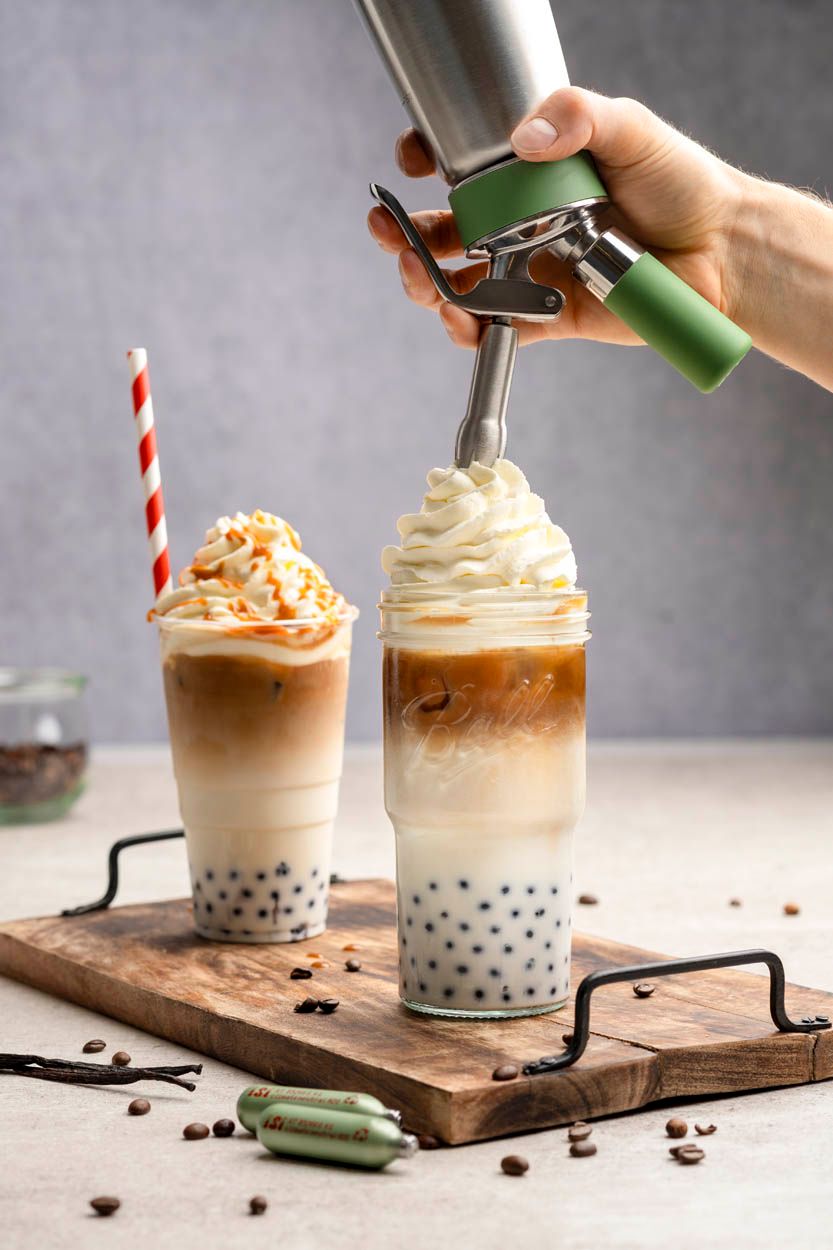 Jemand sprüht mit einem iSi Green Whip Sahneschaum auf einen von zwei Iced Bubble Coffees. Daneben liegen zwei grüne iSi Sahnekapseln. 