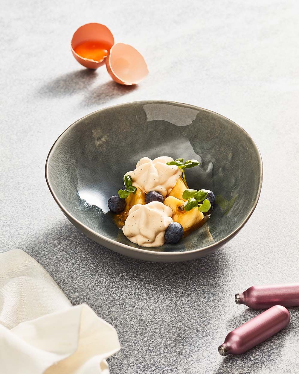 Eine Schüssel mit Creme Brulee Schaum, daneben ein Ei und zwei iSi Charge Kapseln. 