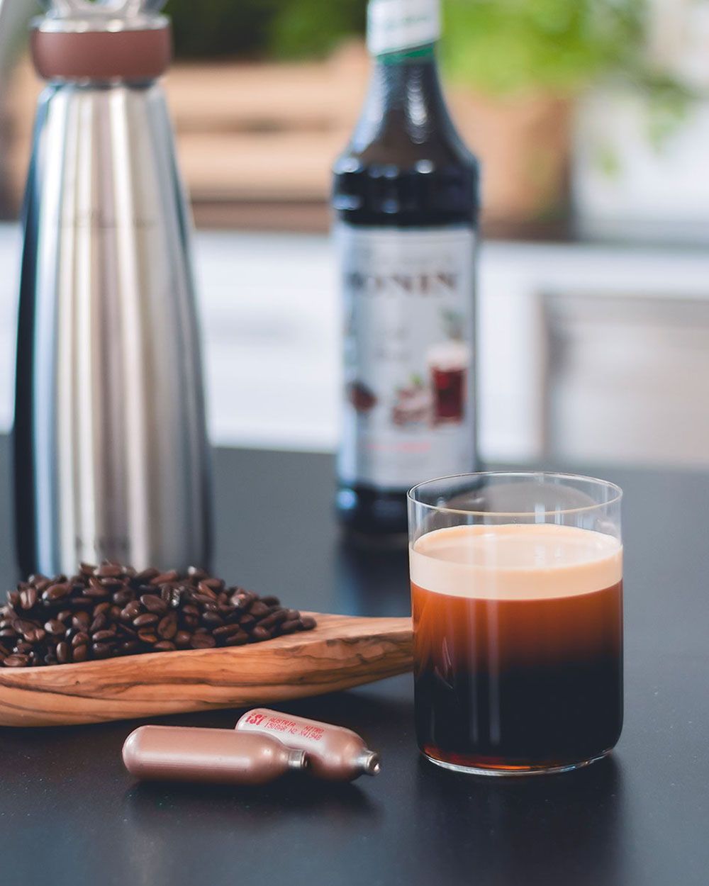 Ein Glas mit Negroni, daneben Kaffeebohnen, iSi Charge Kapseln,  Monin Sirup und ein iSi Nitro. 