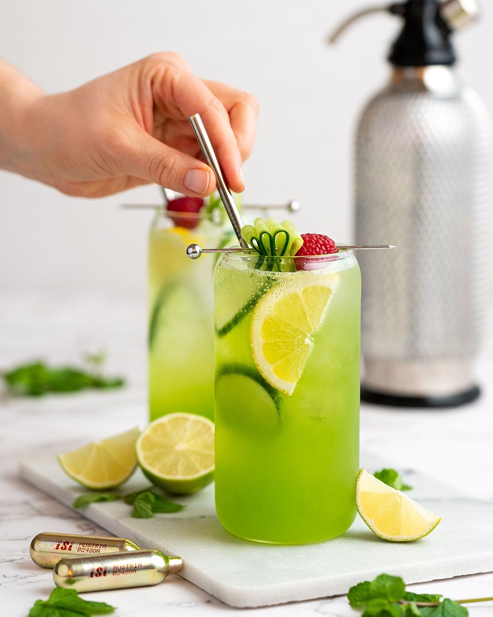 Jemand rührt in einem Glas mit Nitro Gurken Gin um, daneben iSi Charge Kapseln, Limetten und ein iSi Soda Maker. 