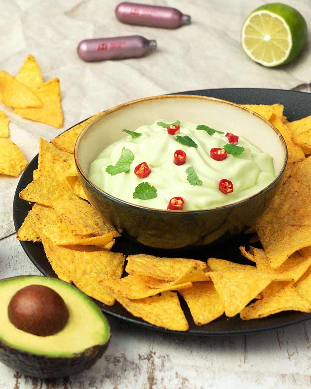 Eine Schale mit cremigem Avocado-Dip, garniert mit Koriander und roten Chilischeiben, steht in der Mitte eines Tellers mit knusprigen Tortilla-Chips, daneben liegen eine Avocadohälfte und eine Limette.