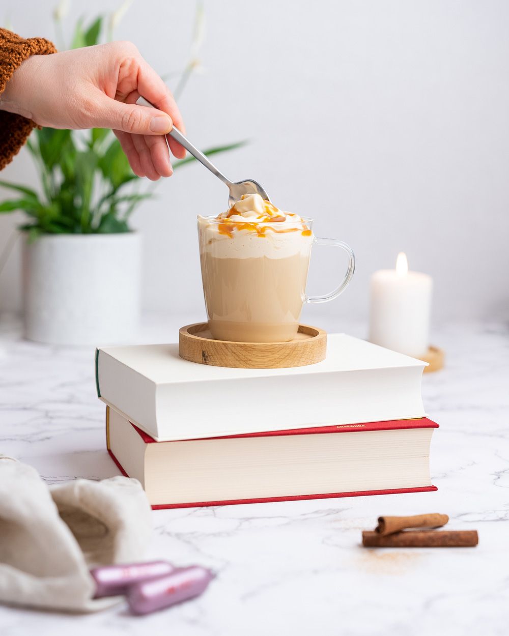 Jemand sticht mit einem Löffel in ein Kaffeegetränk mit Schaum, das auf einem Stapel Bücher steht. Davor liegen Zimtstangen und iSi Charge Kapseln. 