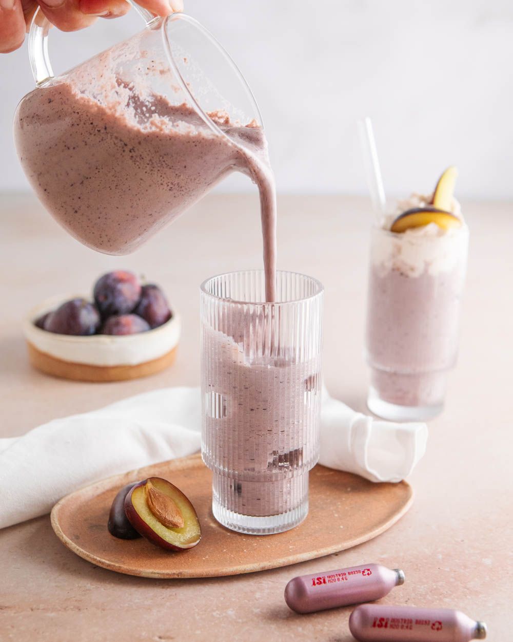 Jemand gießt ein Pflaumen-Milch-Getränk in ein Glas, davor liegen iSi Charge Kapseln. 