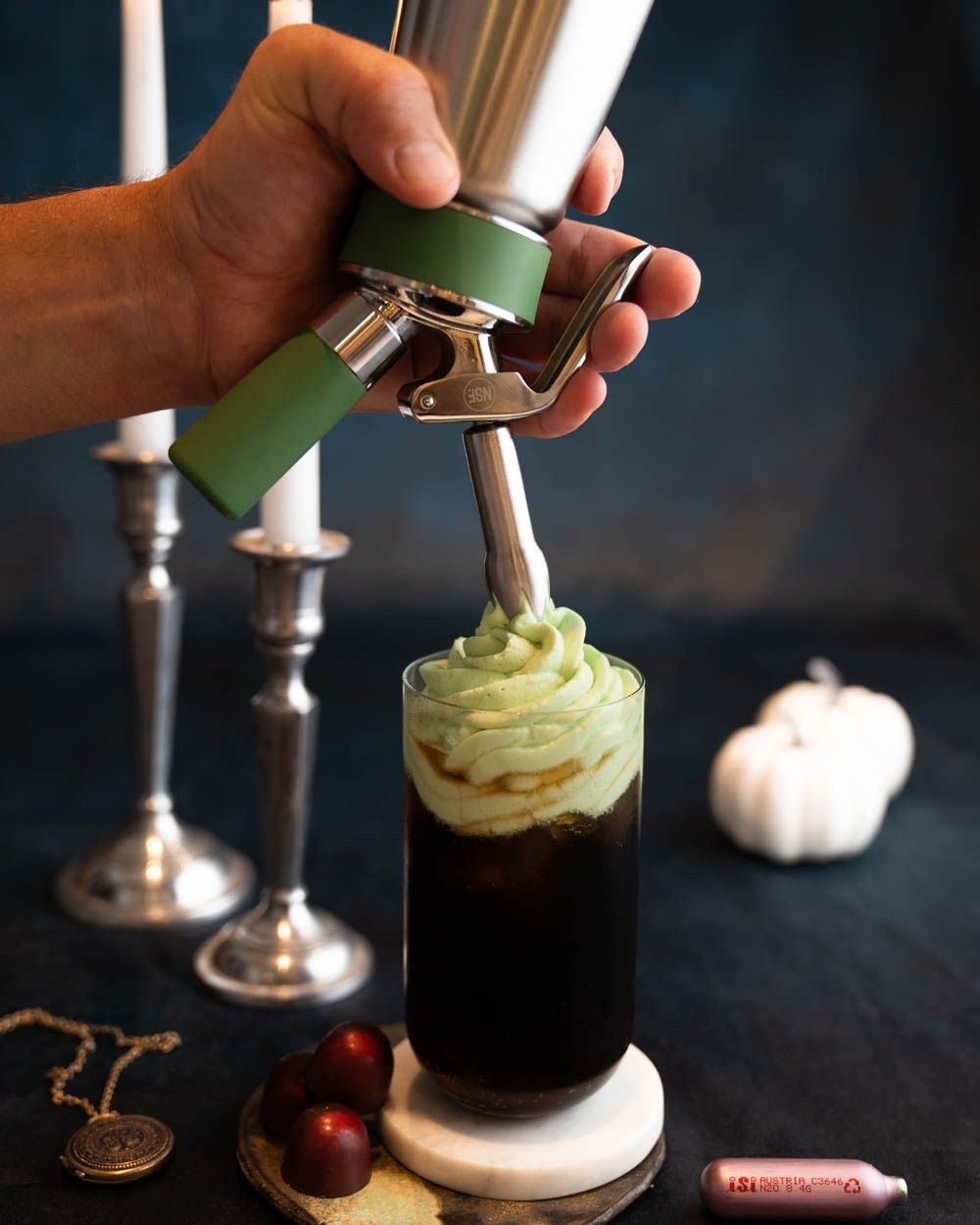 Jemand sprüht mit einem iSi Gourmet Whip Sour Apple Topping auf ein Kaffeegetränk. Im Hintergrund Kürbisse, Kerzenständer und iSi Charge Kapseln. 