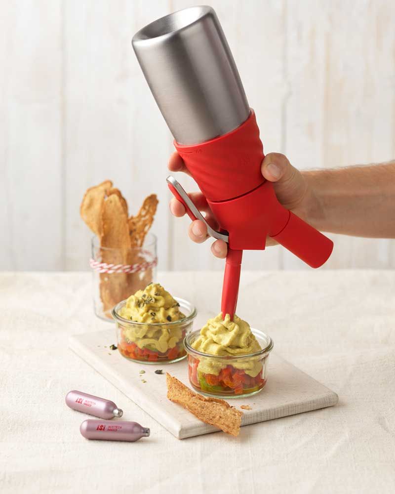 Jemand sprüht mit einem iSi Creative Whip Linsen-Espuma in eine Schüssel, daneben eine weitere Schüssel und zwei iSi Charge Kapseln. Im Hintergrund ein Glas mit Brot-Sticks. 