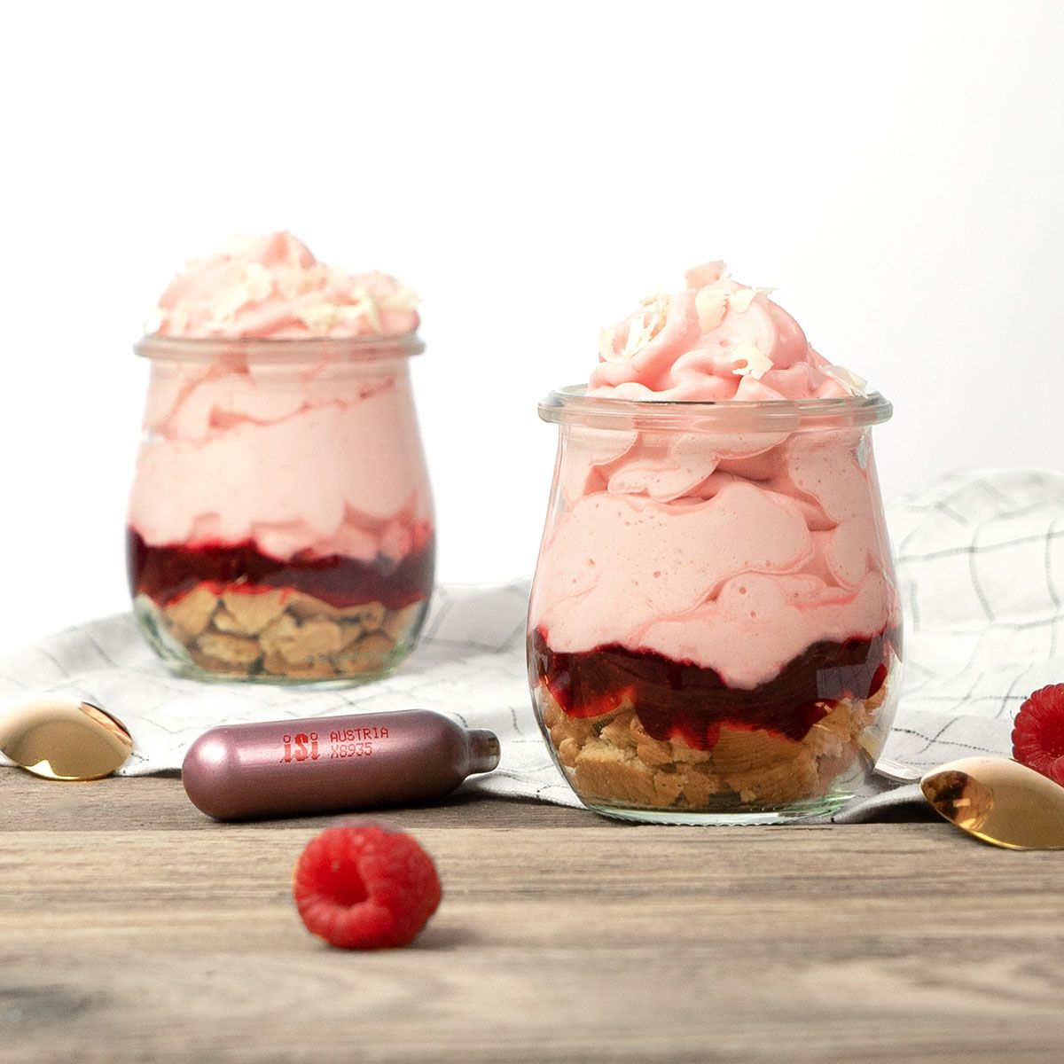 Zwei Gläser mit einem Dessert mit Himbeer-Preiselbeer-Espuma. Daneben Himbeeren und  eine iSi Charge Kapsel.