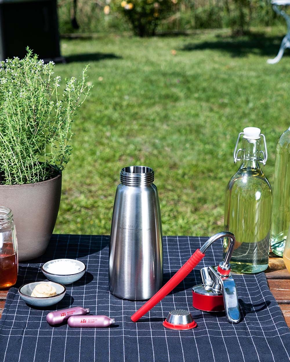 Ein Tisch im freien auf dem ein iSi Gourmet Whip, eine Glasflasche, Gewürze und zwei iSi Charge Kapseln liegen. 