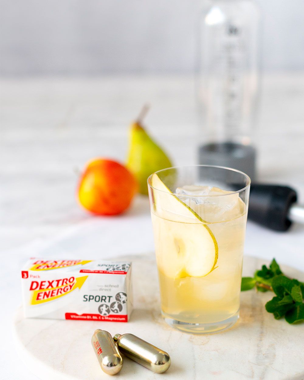 Ein Glas mit einem Birne-Energie-Getränk, daneben iSi Charge Kapseln, Dextro Energy und Obst. 