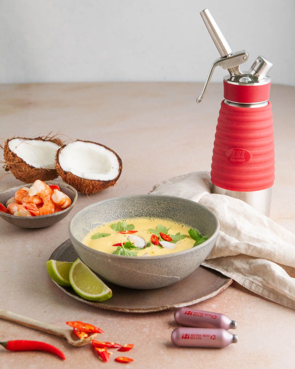 Ein Teller mit thailändischer Kokos-Suppe, daneben Kokosnüsse, Limetten, Gemüse, iSi Charge Kapseln und ein iSi Gourmet Whip. 