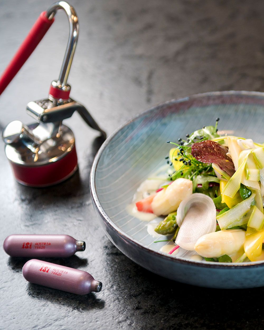 Ein Teller mit Gemüse, daneben ein iSi Gourmet Whip Aufsatz und iSi Charge Kapseln. 