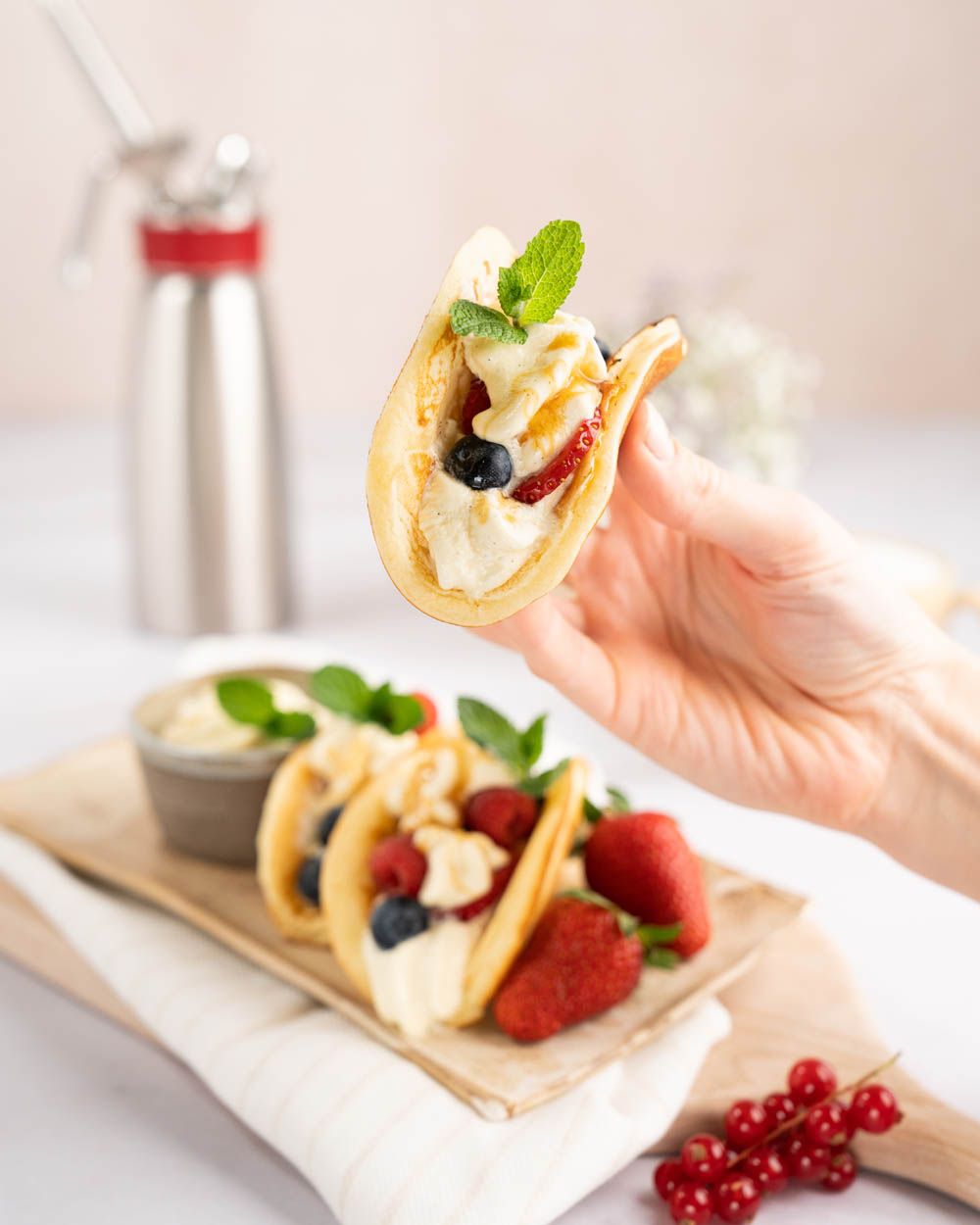 Jemand hält einen Dessert-Taco mit Beeren und Vanille-Mascarpone Creme, im Hintergrund weitere Tacos und ein iSi Gourmet Whip.